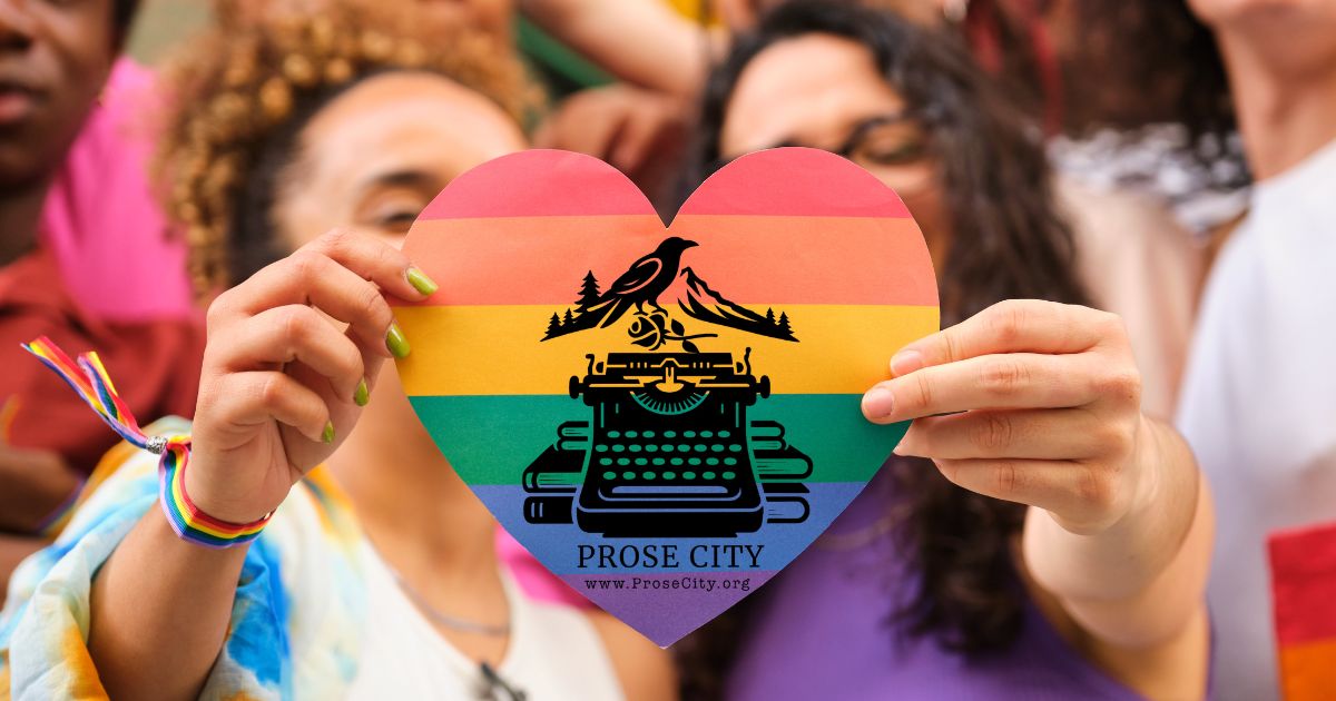 pride-in-prose-city A heart-shaped rainbow flag cutout held up by two people in a crowd. The center of the heart features a black logo of a typewriter, books, and a crow, with the text "PROSE CITY www.ProseCity.org".