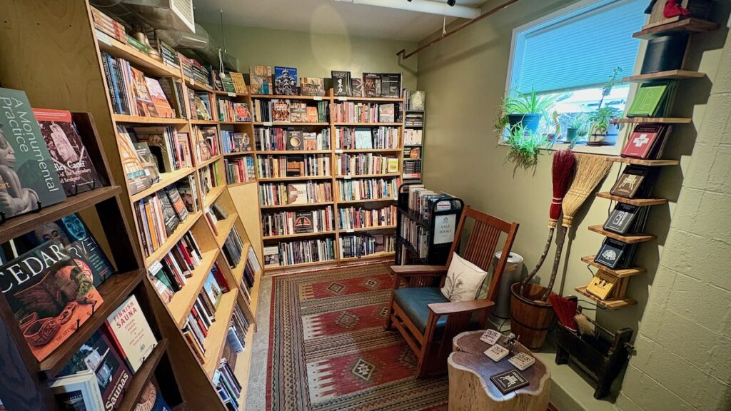 The photograph shows a small, inviting room dedicated to books, many of which appear to be about woodworking, nature, and crafts. On the left and back walls, tall, light-wood bookshelves are tightly packed, with several titles displayed face-out. A patterned red, beige, and blue geometric rug covers the floor. On the right side of the room, a wooden armchair with a blue seat cushion and a white patterned throw pillow sits next to a small side table made from a thick, live-edge tree stump. Behind the chair, a small black rolling cart is labeled 