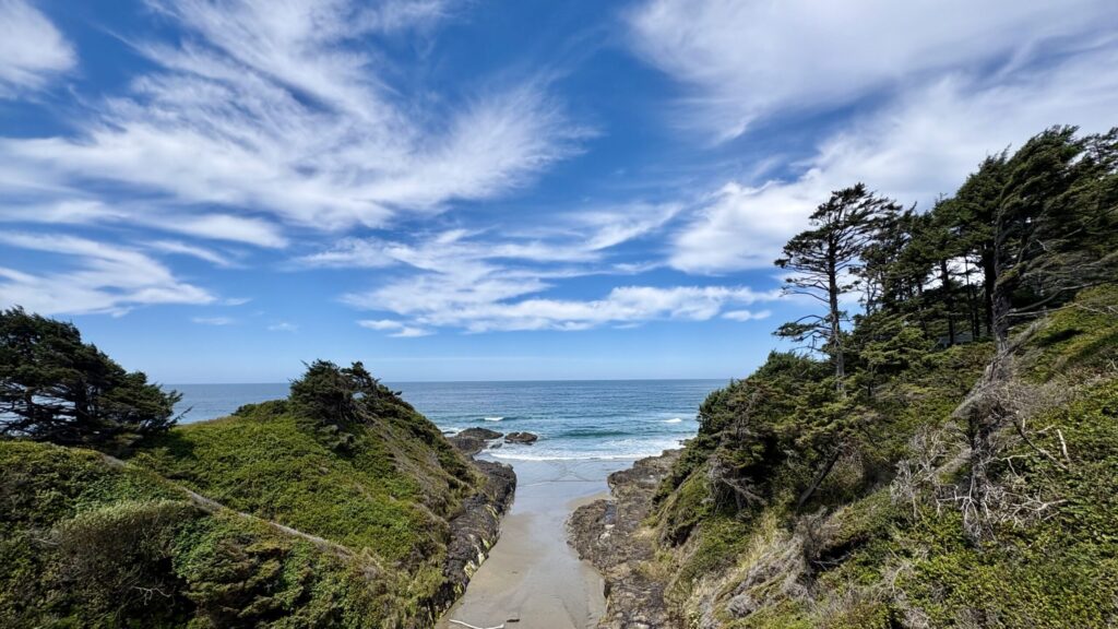 A narrow sandy beach inlet framed by green, tree-covered cliffs leading to the ocean under a bright blue sky with sweeping white clouds.