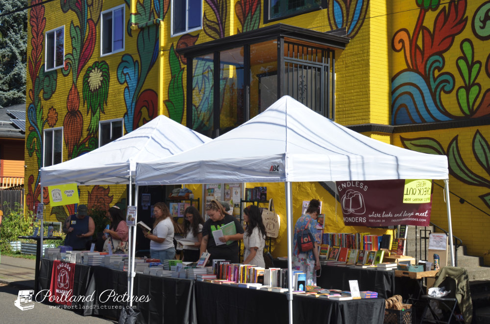 An outdoor book display featuring two white pop-up canopy tents set up along a sidewalk. Beneath the tents, long tables draped in black cloth are covered with arranged stacks and rows of books. Several people stand behind the tables browsing through books. A maroon banner hanging under the canopy reads "ENDLESS WONDERS: where books and beings meet". In the background is a vibrant two-story yellow building adorned with a large, colorful floral and leafy mural. A "Portland Pictures" logo watermark is in the lower-left corner.