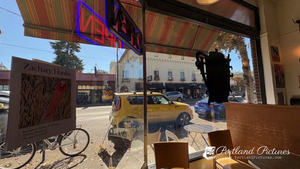 A view looking out from inside a coffee shop through large glass windows onto a sunny street scene. In the foreground inside the shop, wooden chairs, a table, and a booth seat are visible beneath a red, yellow, and green striped awning. A glowing neon "OPEN" sign hangs reversed above. A black outline silhouette of a coffee pot is on the right pane of glass.
On the left window pane, a poster for an art exhibition reads "Zachary Hanks" with event details. Outside on the sidewalk, a white bicycle is parked on the left. Across the street, a bright yellow compact car and a blue sedan are parked along the curb, with multi-story brick and stucco buildings, shop fronts, and green trees lining the street under a clear blue sky. A "Portland Pictures" watermark appears in the bottom-right corner.