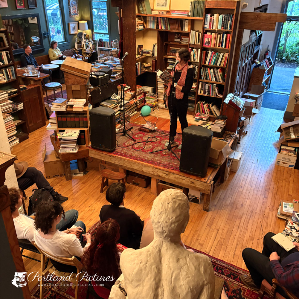 A high-angle, overhead view of a cozy, crowded bookstore during a live reading event. On a low wooden stage covered with a patterned red Persian rug, a person wearing a dark top, dark pants, and a pink scarf stands holding an open book and speaking into a microphone on a stand. Black speakers flank either side of the stage.
The room is lined with tall wooden bookshelves packed with books, along with stacks of books on the floor and tables. In the foreground, the backs of several audience members seated on chairs and the floor face the stage. A large, light-colored plaster bust or sculpture stands prominently in the bottom center foreground. In the upper left background, additional patrons sit at tables near a window. A "Portland Pictures" watermark appears in the bottom-left corner.