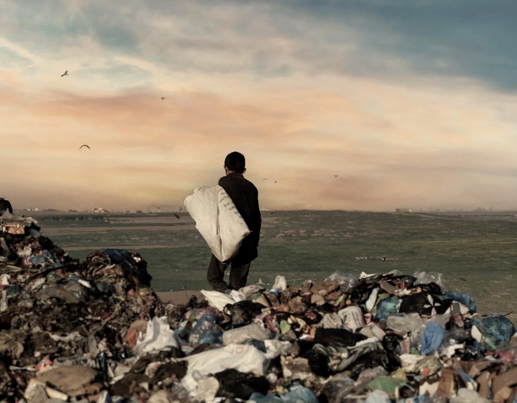 A young person in dark clothing is photographed from behind, standing at the edge of a landfill with a large, worn white sack slung over their shoulder. Printed text, partly legible, is visible on the sack. In the foreground, heaps of mixed waste fill the bottom of the frame: plastic bags in white, blue, and black, torn packaging, cardboard, and organic debris. Beyond the trash, a wide, flat green plain stretches toward a hazy skyline with a few low buildings. Several birds are in flight, and a small group of animals, possibly sheep or goats, grazes in the distance. The sky is dramatic, with warm orange and peach clouds fading into dusty blue and teal at the top.