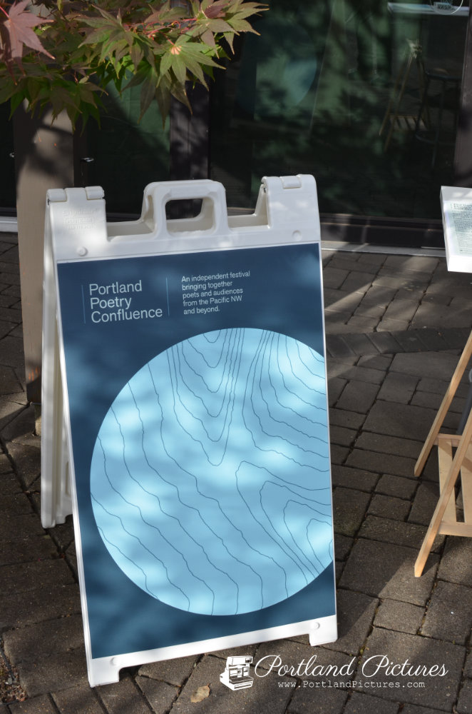A white plastic A-frame sign rests on a paved stone walkway outdoors. The sign features a dark blue face displaying white text that reads, "Portland Poetry Confluence," alongside the tagline, "An independent festival bringing together poets and audiences from the Pacific NW and beyond." Below the text is a large light-blue graphic circle filled with dark contour lines resembling a topographic map. Sunlight casts tree branch shadows across the sign, and Japanese maple leaves hang over the top-left corner. A "Portland Pictures" watermark appears in the bottom-right corner.