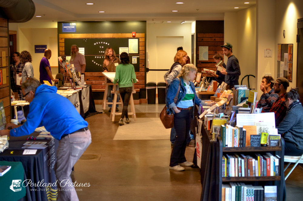 A wide indoor room filled with people browsing vendor tables at a book fair or community event. In the foreground on the left, a person in a bright blue hoodie bends over a black tablecloth-covered display table filled with papers and items. On the right, a multi-tiered wooden shelf and tables display rows of books where several attendees are standing and browsing. In the background, a wooden-paneled wall displays a black circle sign reading "WELCOME TO THE POETRY CONFLUENCE". The room features warm overhead lighting and polished concrete floors. A "Portland Pictures" watermark appears in the bottom-left corner.