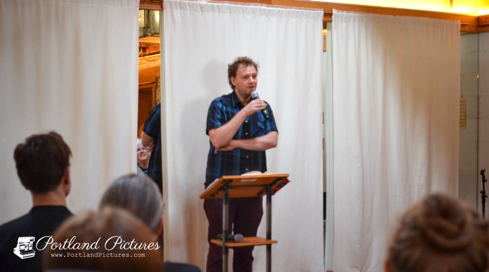 A person with short curly brown hair and a blue-and-black plaid short-sleeved shirt stands behind a tall wooden podium or lectern. They hold a microphone in their left hand near their chin, with their right arm crossed over their stomach as they address the room. Behind them is a background of white hanging curtains divided into sections. In the foreground, the backs of several audience members' heads are visible, facing toward the speaker. A logo watermark reading "Portland Pictures" appears in the bottom-left corner.