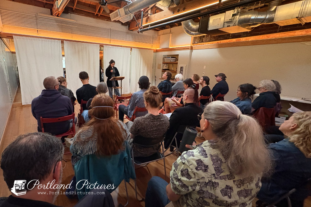 A wide shot from behind an audience seated in dark and red folding chairs facing the front of an indoor event space. At the front of the room, a person stands behind a wooden podium speaking into a microphone in front of hanging white curtains. The room features warm wood flooring, high exposed ceilings with wooden beams, large metal air ducts, and warm ambient lighting along the top of the walls. The audience members are dressed casually and focused toward the stage. A "Portland Pictures" watermark appears in the bottom-left corner.