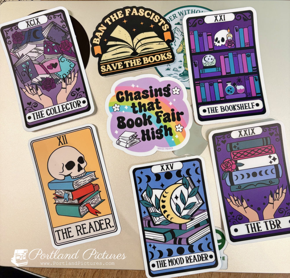 A group of colorful, book-themed vinyl stickers arranged on a silver laptop lid.
The collection includes five tarot card-style stickers:
"THE COLLECTOR" (top left, Roman numeral XCIX): Features hands holding a sprawling stack of open, glowing books.
"THE BOOKSHELF" (top right, Roman numeral XXI): Depicts bookshelves filled with colorful books, a skull, a candle, and potions.
"THE READER" (bottom left, Roman numeral XII): Shows a human skull sitting atop a stack of colorful books.
"THE MOOD READER" (bottom center, Roman numeral XXV): Features open books beneath a crescent moon surrounded by moon phases.
"THE TBR" (bottom right, Roman numeral XXIX): Displays hands balancing a tall stack of books.
Two non-tarot stickers sit in the center:
Top Center: An arch-shaped black and orange sticker showing an open book that reads "BAN THE FASCISTS SAVE THE BOOKS".
Center: A cloud-shaped sticker with a rainbow and flowers that reads "Chasing that Book Fair High" above a small stack of books.
A "Portland Pictures" watermark appears in the bottom-left corner.