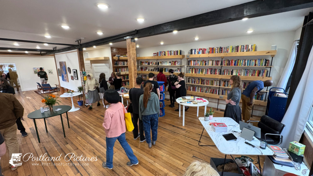 A high-angle view looking down into a spacious, well-lit bookstore and art space. Warm hardwood floors stretch across the room beneath dark structural ceiling beams and recessed lighting. Along the right wall, long wooden shelves mounted on black brackets are filled with colorful books. Several visitors walk around and browse the shelves, while small round white and green tables with books and floral displays sit in the open floor space. In the foreground on the right, a white marble-patterned desk holds a laptop, card reader, and checkout supplies. Artwork is mounted on white walls in the background. A "Portland Pictures" watermark appears in the bottom-left corner.