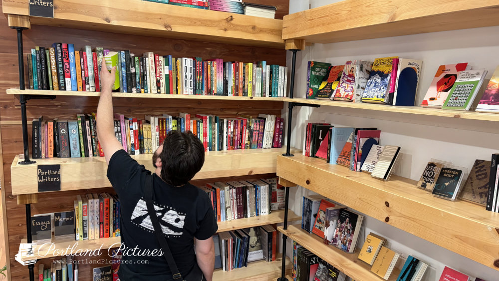 A person wearing a black T-shirt with a graphic on the back and a cross-body bag stands facing a set of light-colored wooden bookshelves. They are reaching their right arm high up to pull a green book off the top shelf. The shelves, constructed with industrial black pipe supports, are filled with rows of books categorized by handwritten labels such as "Portland Writers" and "Essays / Memoir". To the right, another section of shelving displays books face-out, showcasing their cover art. A "Portland Pictures" watermark appears in the bottom-left corner.